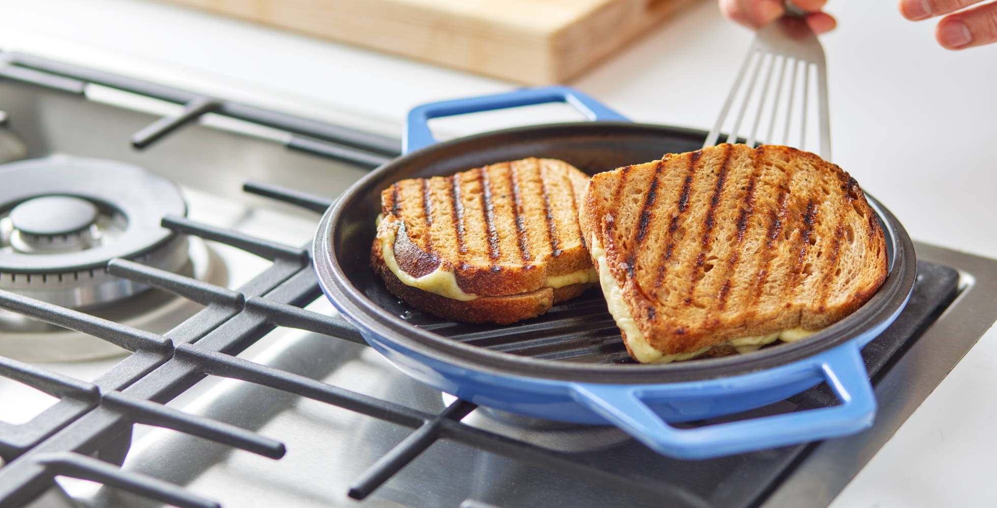 A hand uses a fish spatula to flip a grilled cheese sandwich with clear grill marks in a Blue Grill Lid on a stovetop.