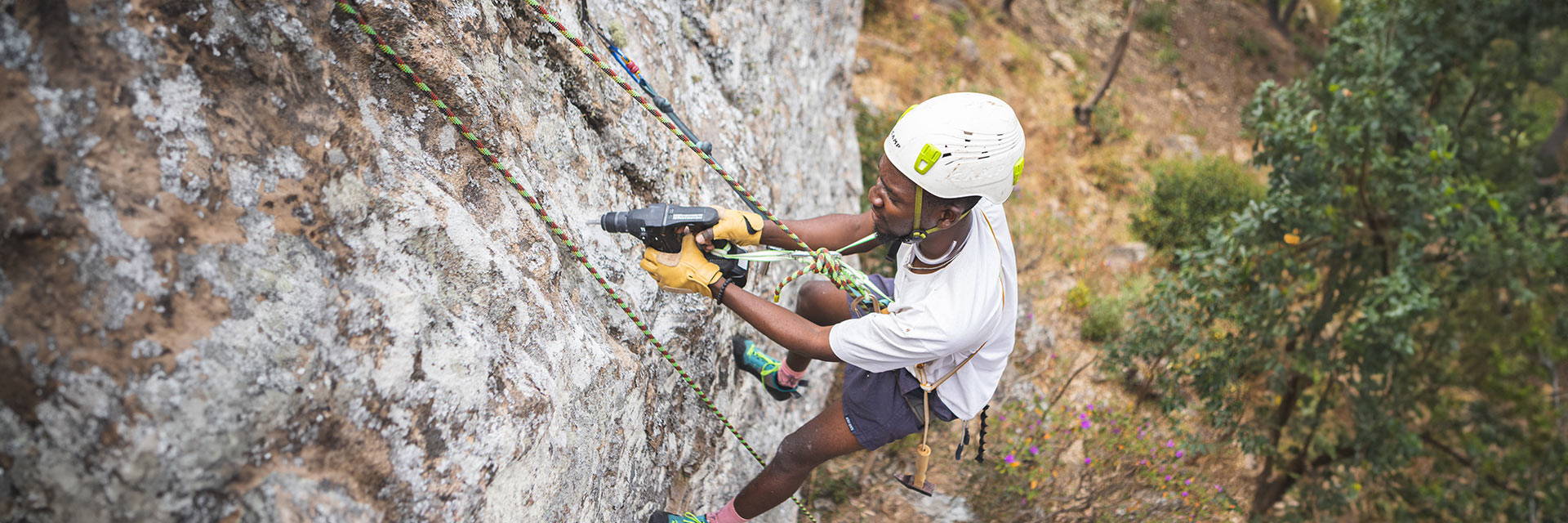 A Malawian climber drills into rock while hanging on a rope to develop a route