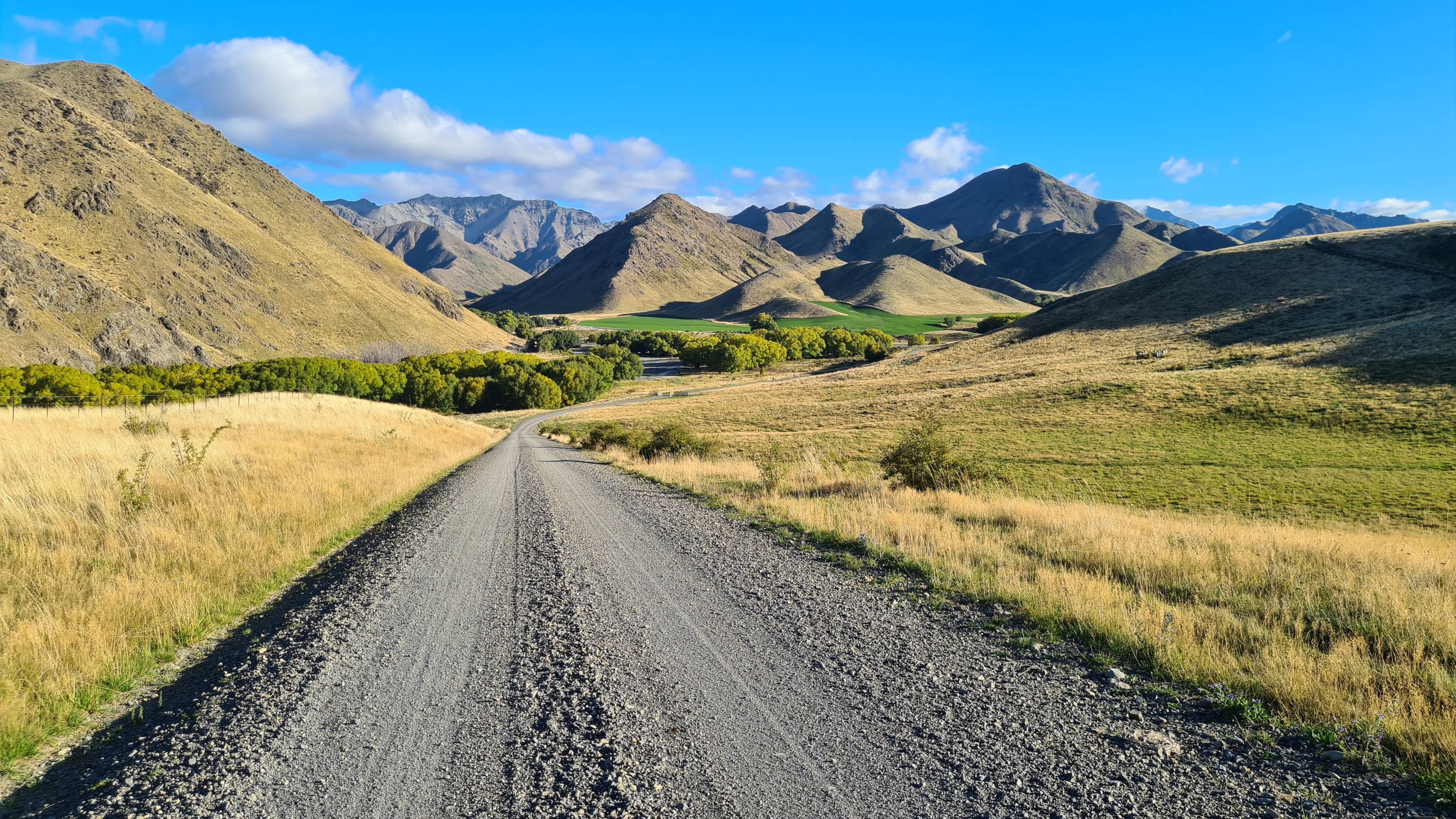 A long stretch of dark gravel road winds towards a mountain range on the horizon.