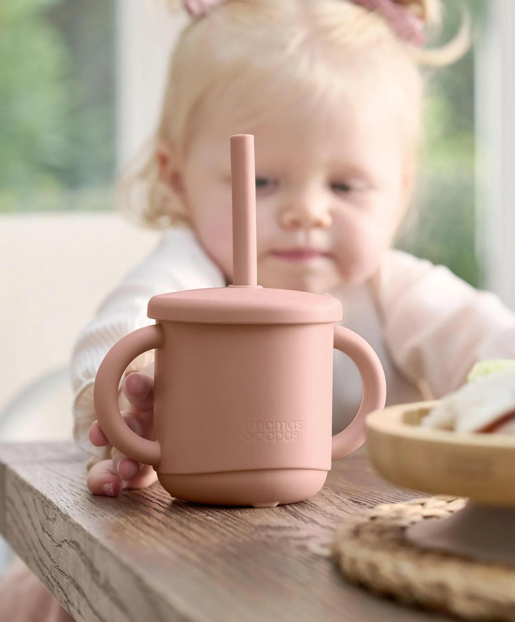 A toddler reaching for a pink sippy cup on a wooden table.