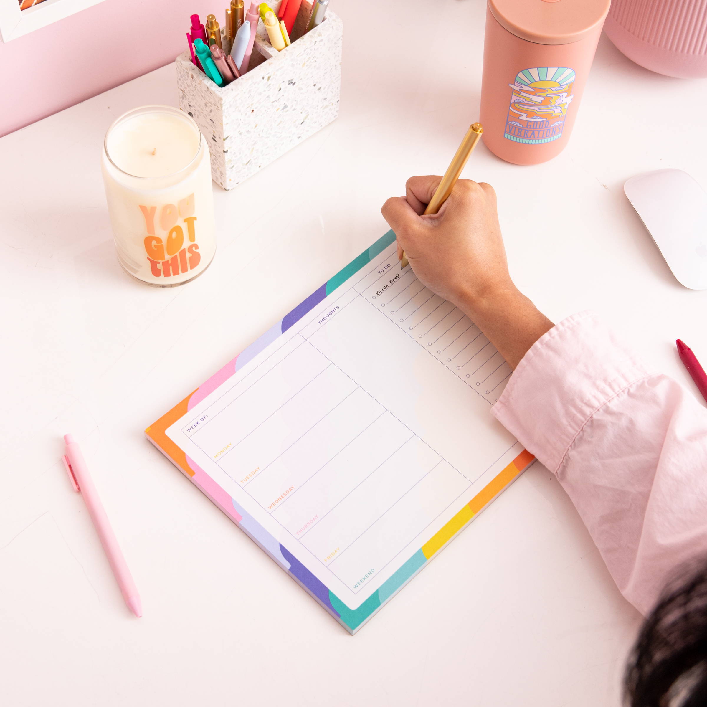 An over view of a person sitting at a desk writing on a weekly desk pad, also know as a Tearaway Notepad. The Notepad has a Monday-Friday  view with a section for thoughts and to-do. On the boarder of the notepad it has a colorful scallop line pattern.  There is also pens,  a candle, and cold cup. 