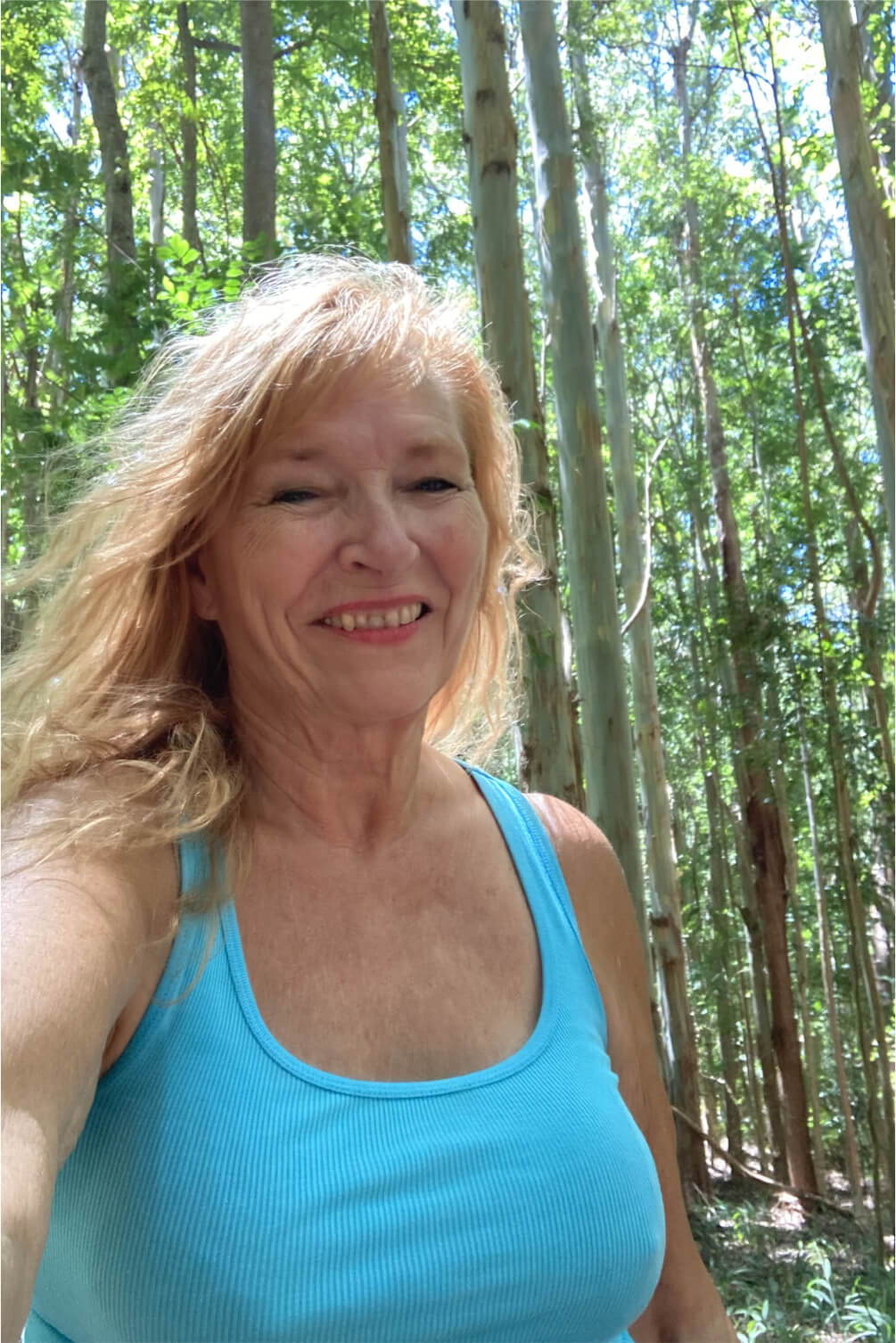 Smiling woman in a blue tank top among tall trees.