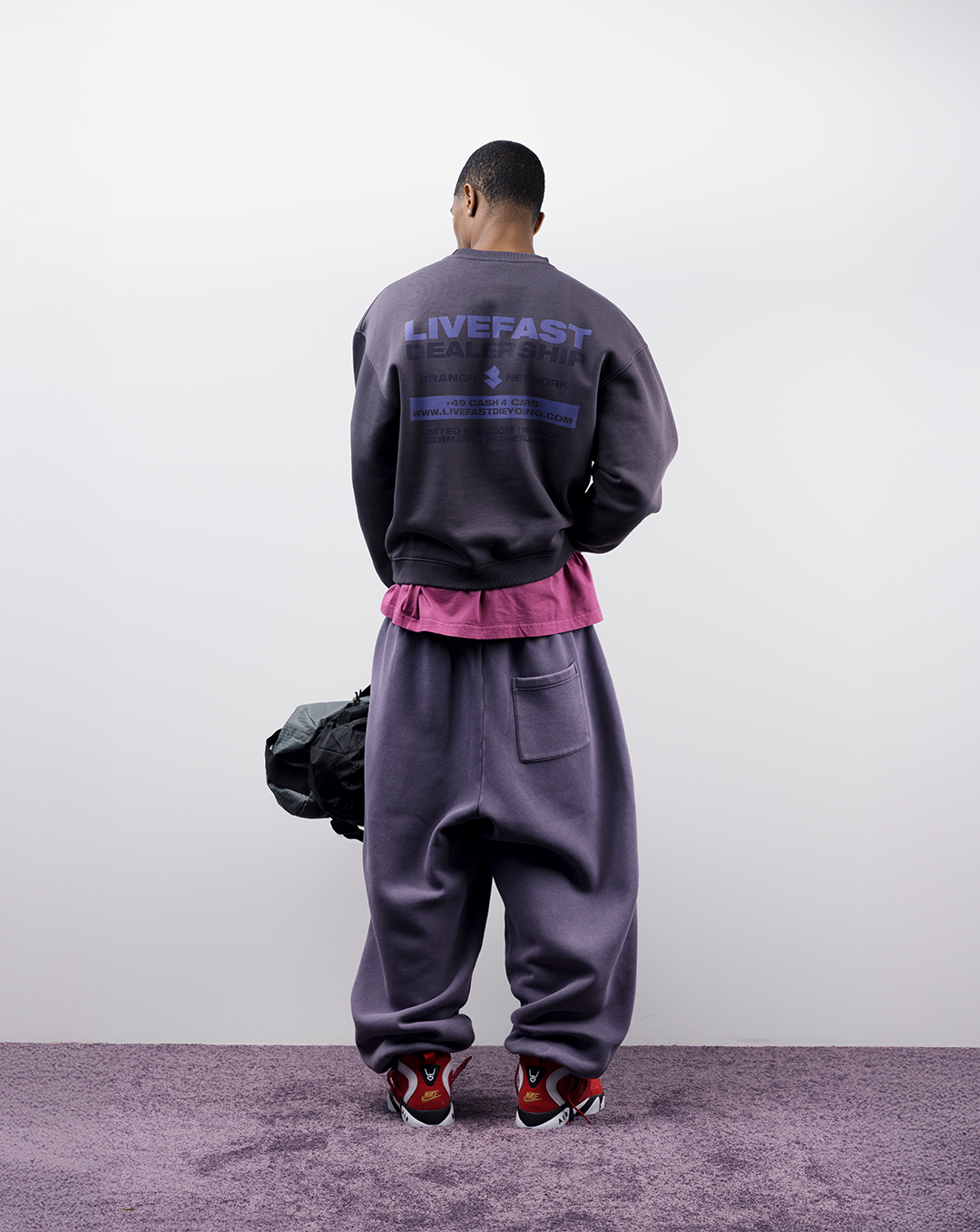 Person wearing oversized purple outfit, facing away, holding a black bag.