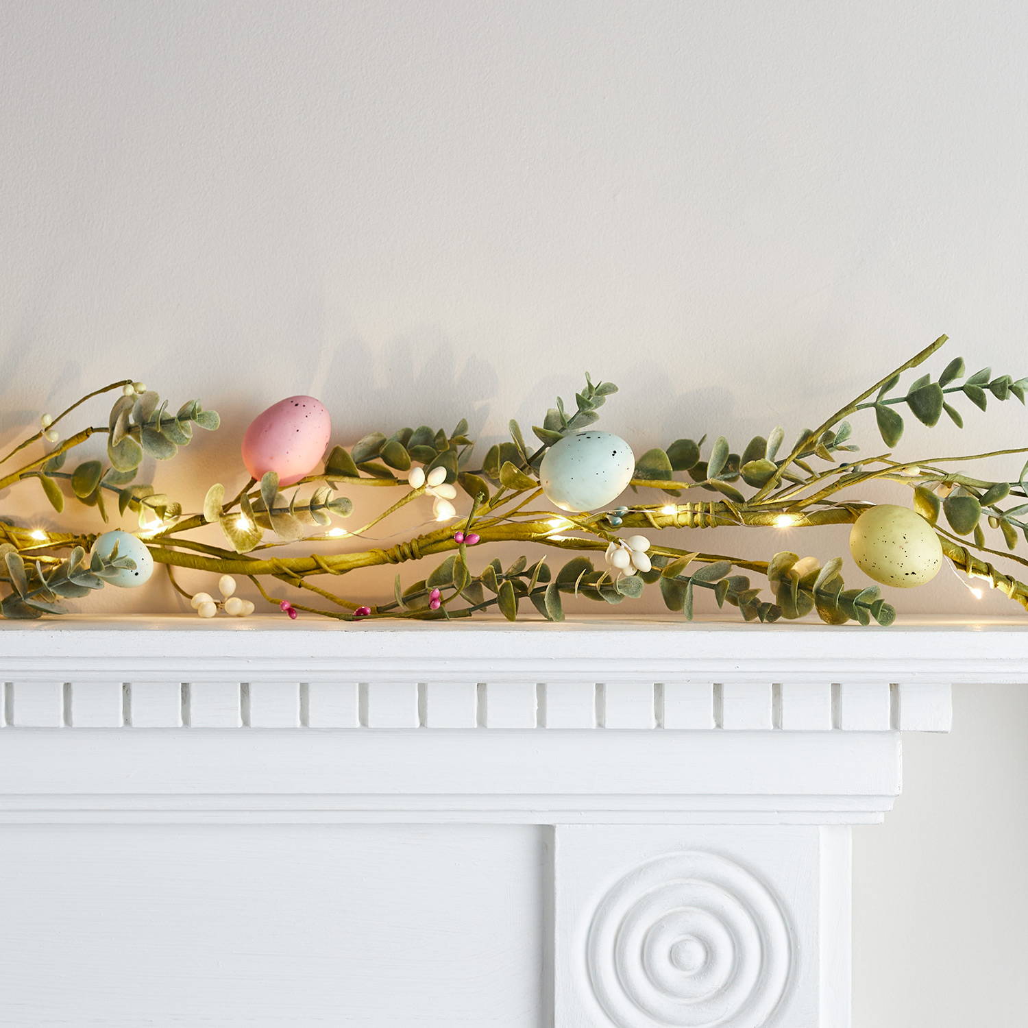An Easter wreath on a mantlepiece with colourful eggs, berries and faux foliage wrapped in micro lights.