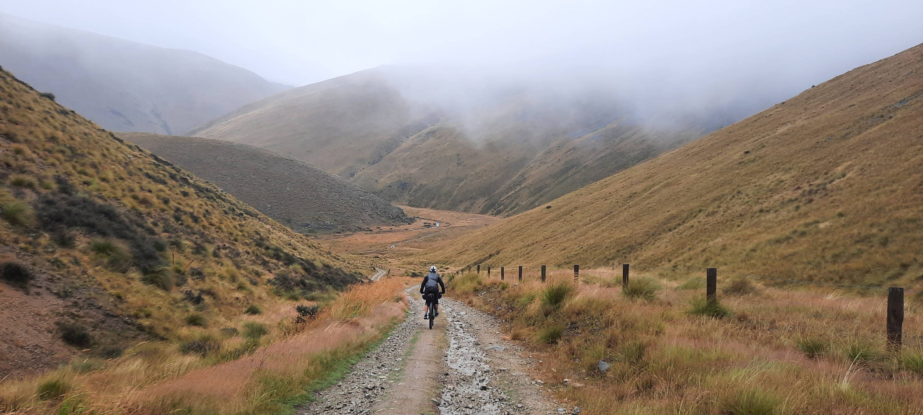 A cyclist rides alone along a double-track gravel road into a foggy hillside.