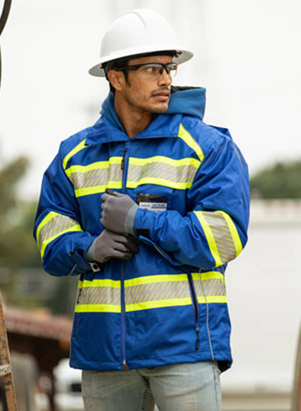 Construction worker wearing blue parka with hi-vis and reflective tape, and white hard hat with black rim safety glasses.