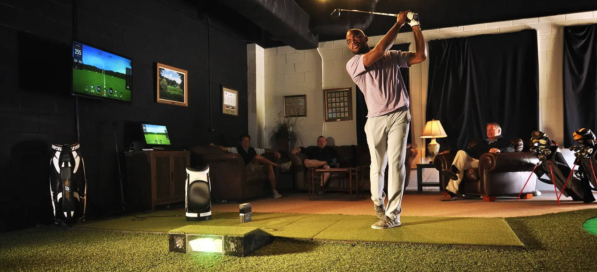 A golfer swinging in a home golf simulator with the SkyTrak and 3 other men sitting and watching in the background
