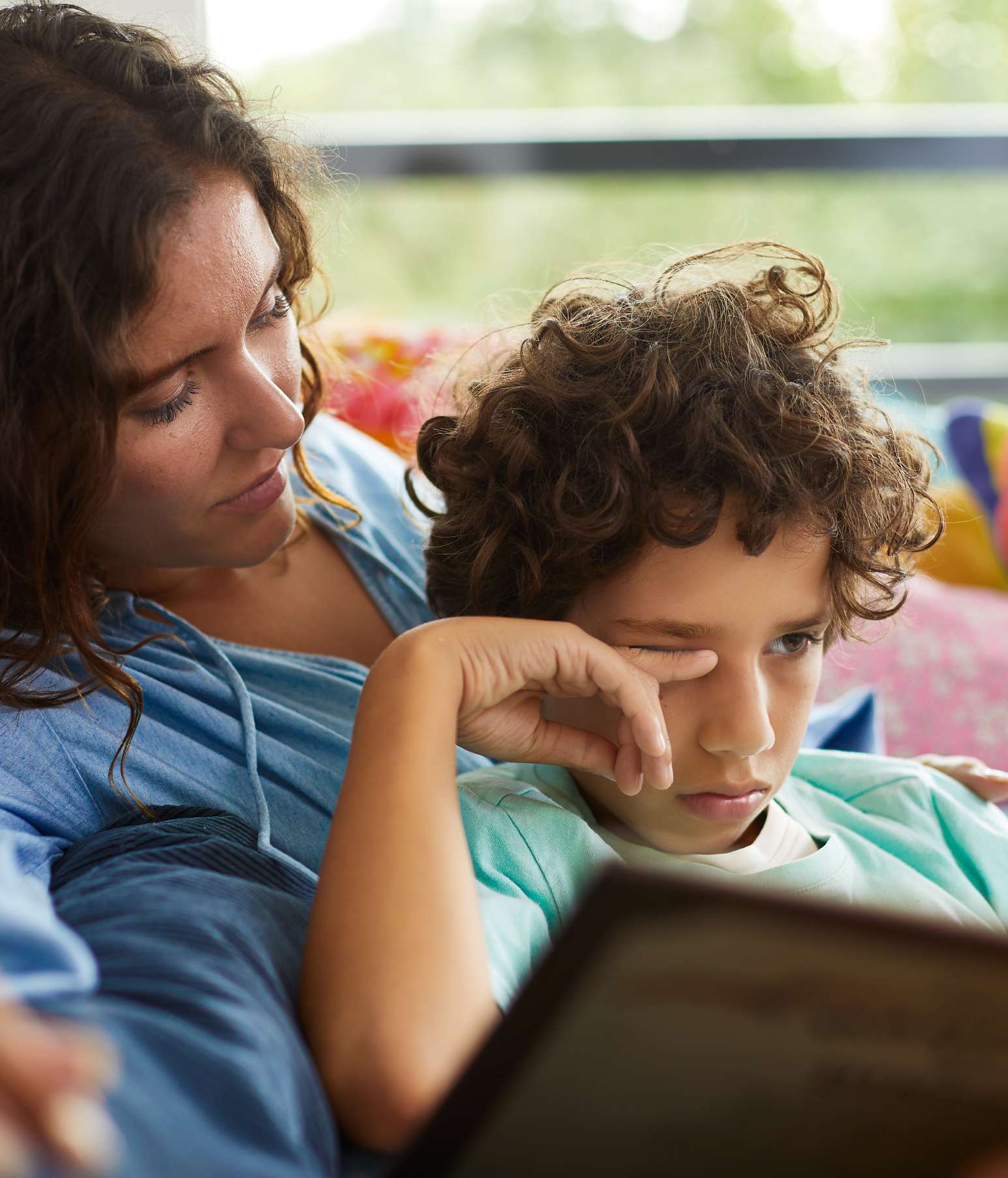 Tired boy rubbing his itchy eye while he sits on the sofa with his mother and a book – his allergies are acting up