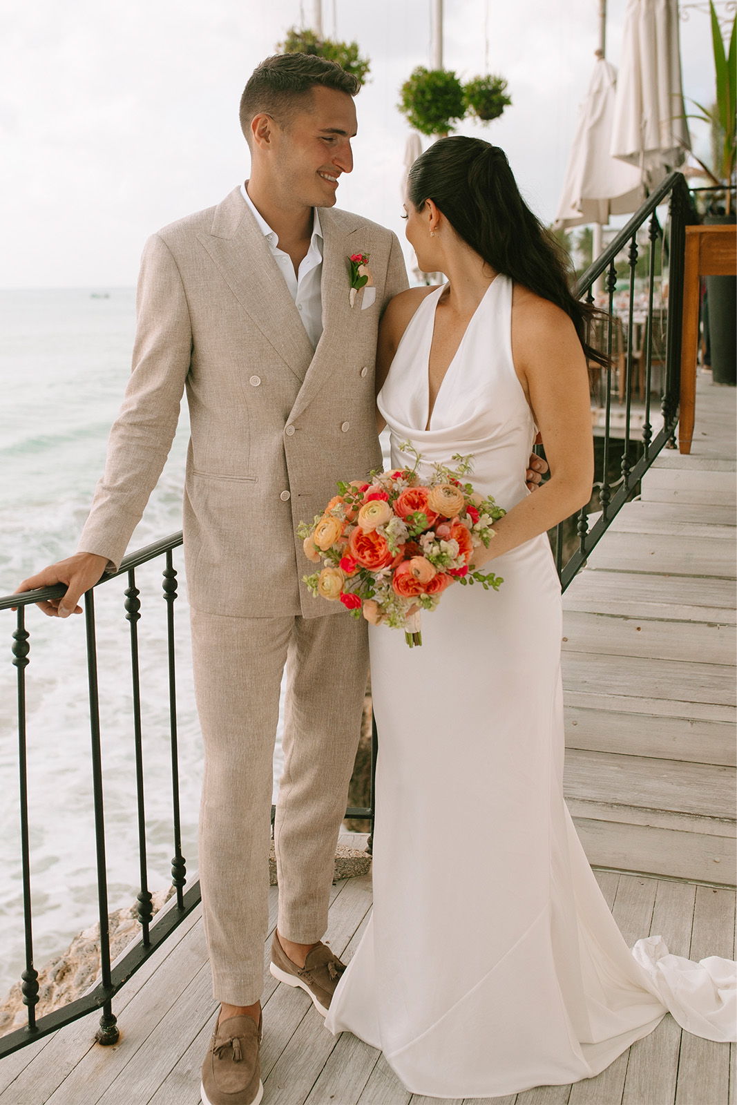 Couple in wedding attire standing by the ocean, smiling and holding a bouquet.