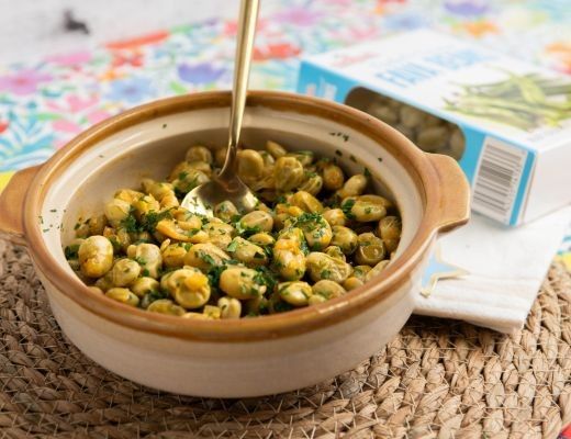 A bowl of cooked legumes with a spoon, accompanied by a box.