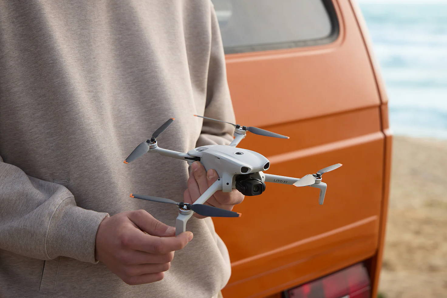 Person holding a DJI Lito X1 next to an orange van by the beach.
