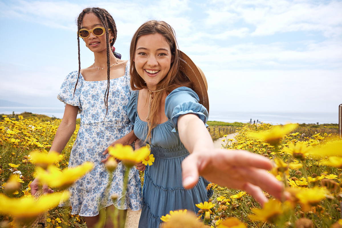 Trixxi sun kissed summer lookbook link, two girls at the beach in field of flowers wearing a cobalt blue dress and a blue floral dress, picking flowers.