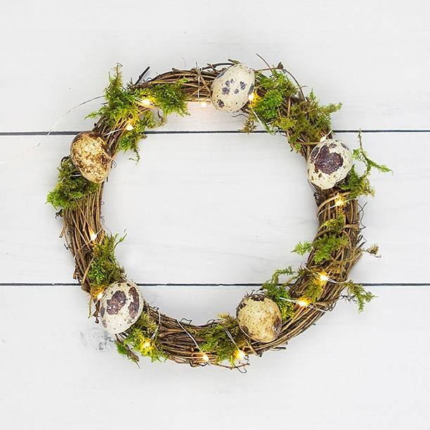 Easter wreath with eggs. foliage and fairy lights in front of a white wooden background.
