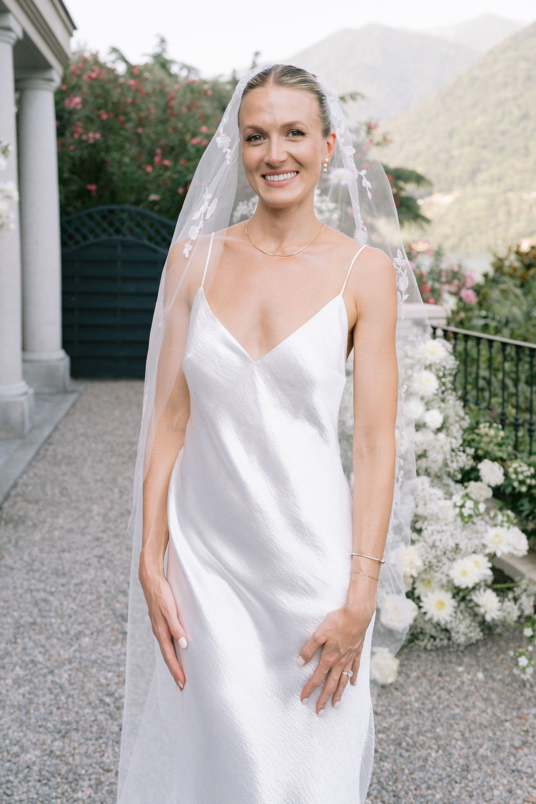 Smiling bride in a white gown with a veil, standing outdoors.
