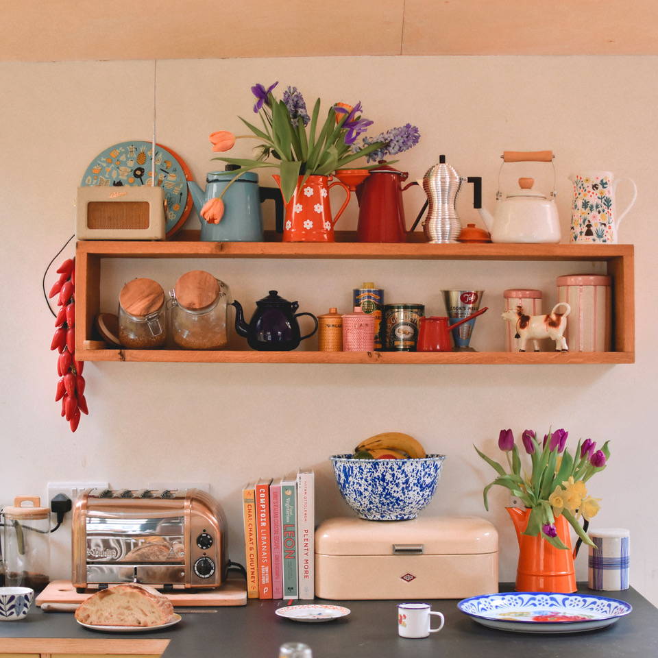 An image of a kitchen surface with toaster and cookbooks and a shelf full of coffee and tea pots and flowers.