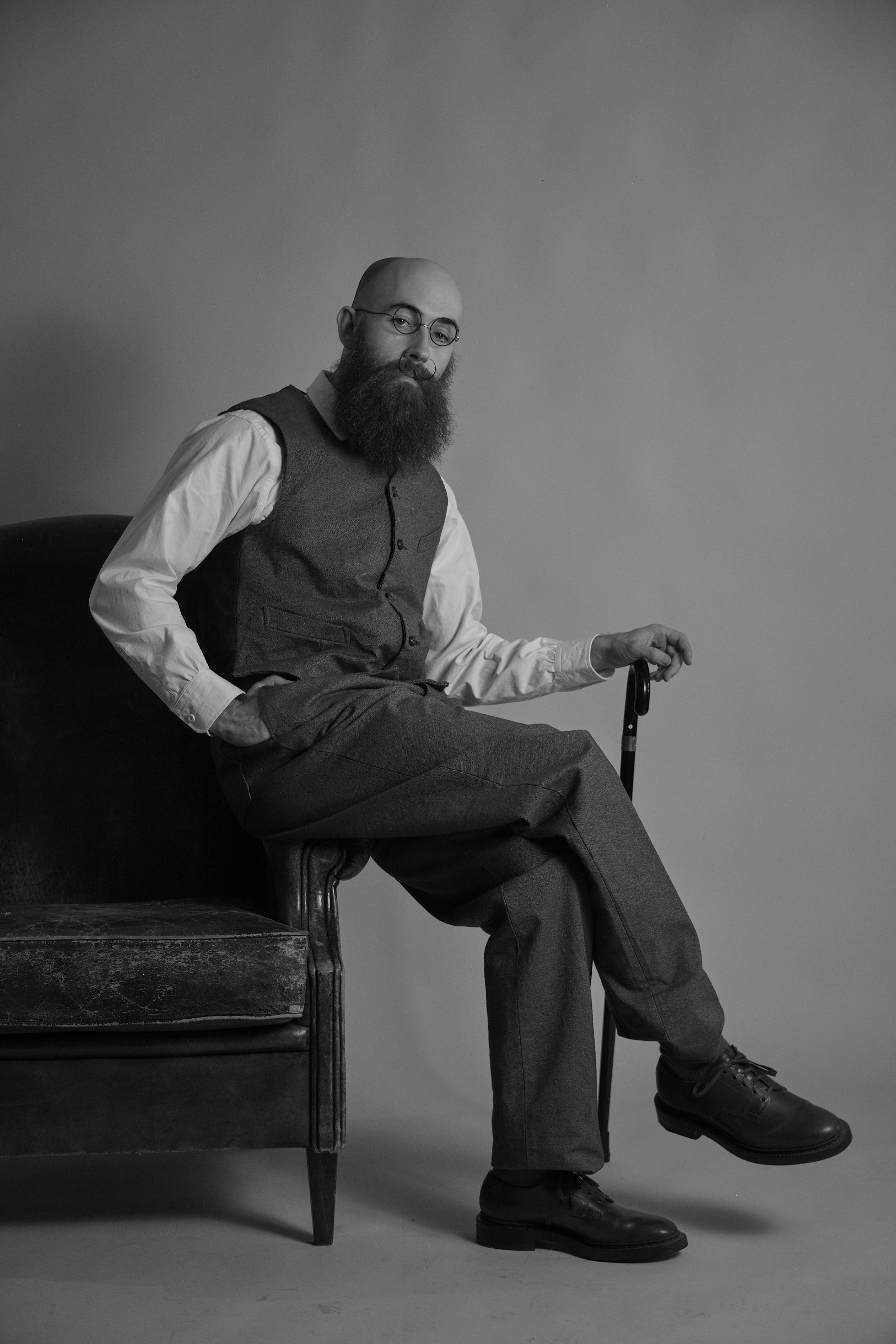 Man with a beard sitting on a chair, wearing a vest and trousers.