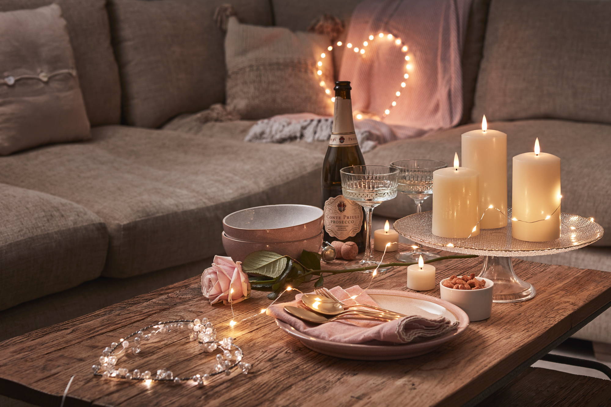 Date night setting with candles, fairy lights and champagne on a table and heart lights on the sofa. 