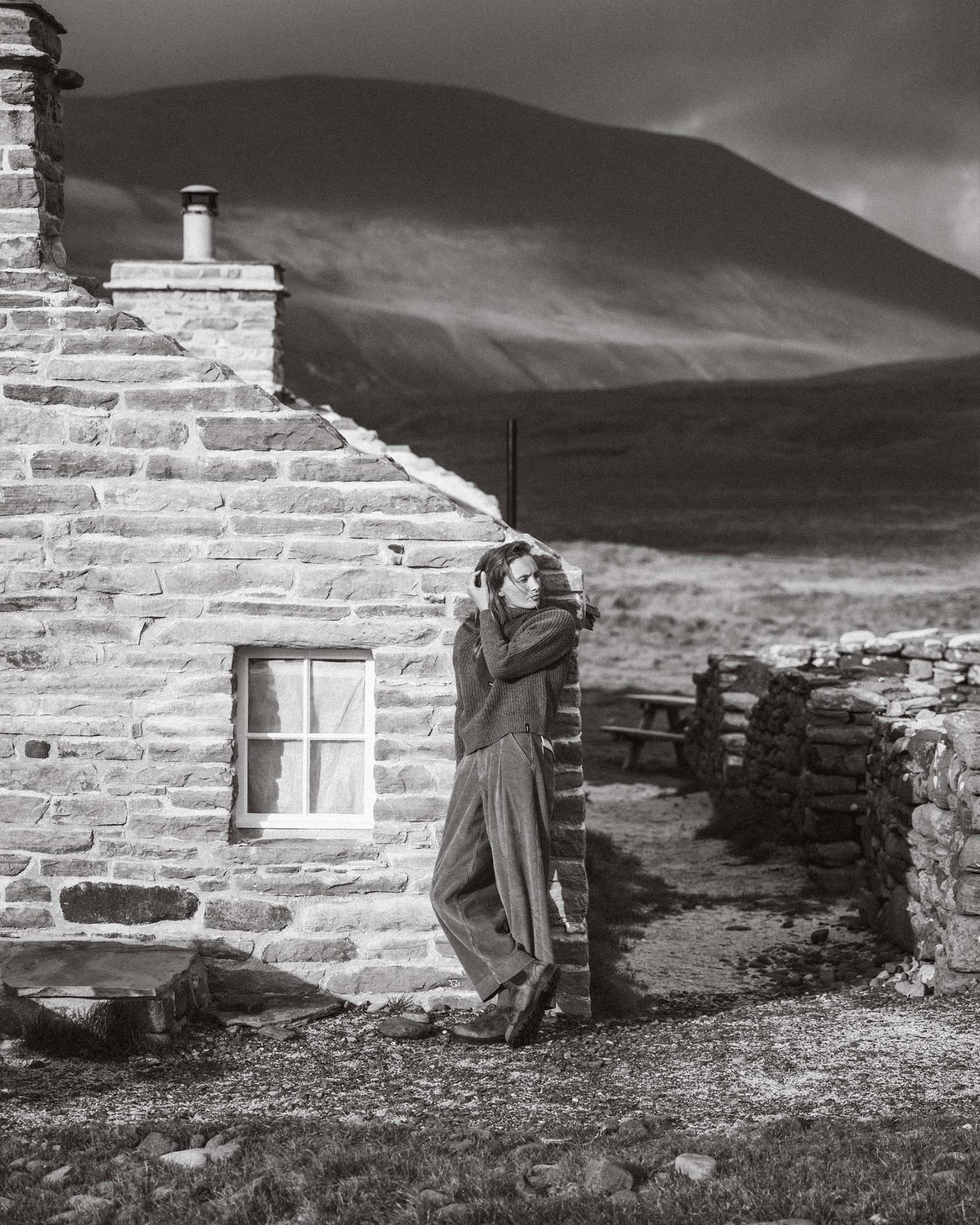 Person standing beside a stone cottage in a mountainous landscape.