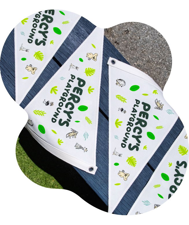 White triangular pennant flags with DTF printed transfers of the Percy's Playground logo and colourful illustrated animals and tropical leaves, laid flat on a blue wooden bench