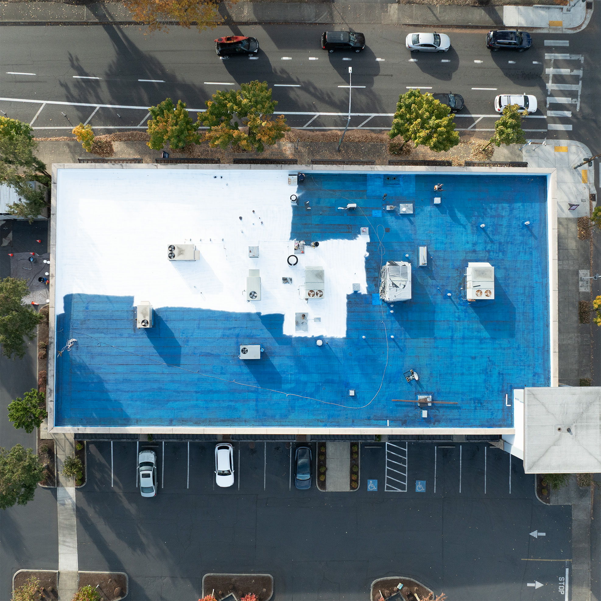 Aerial view of a building roof with blue coating and surrounding parking lot.