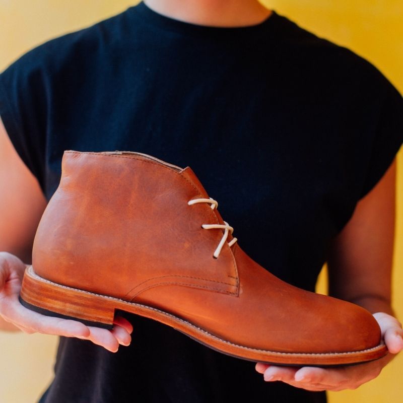 A studio shot of a person wearing a plain black t-shirt holding a single tan or caramel brown leather Adelante Santiago chukka boot horizontally in both hands to display its profile. The lace-up boot features distinct white contrast laces and a cleanly finished stacked leather sole. The background is a solid, vibrant yellow.