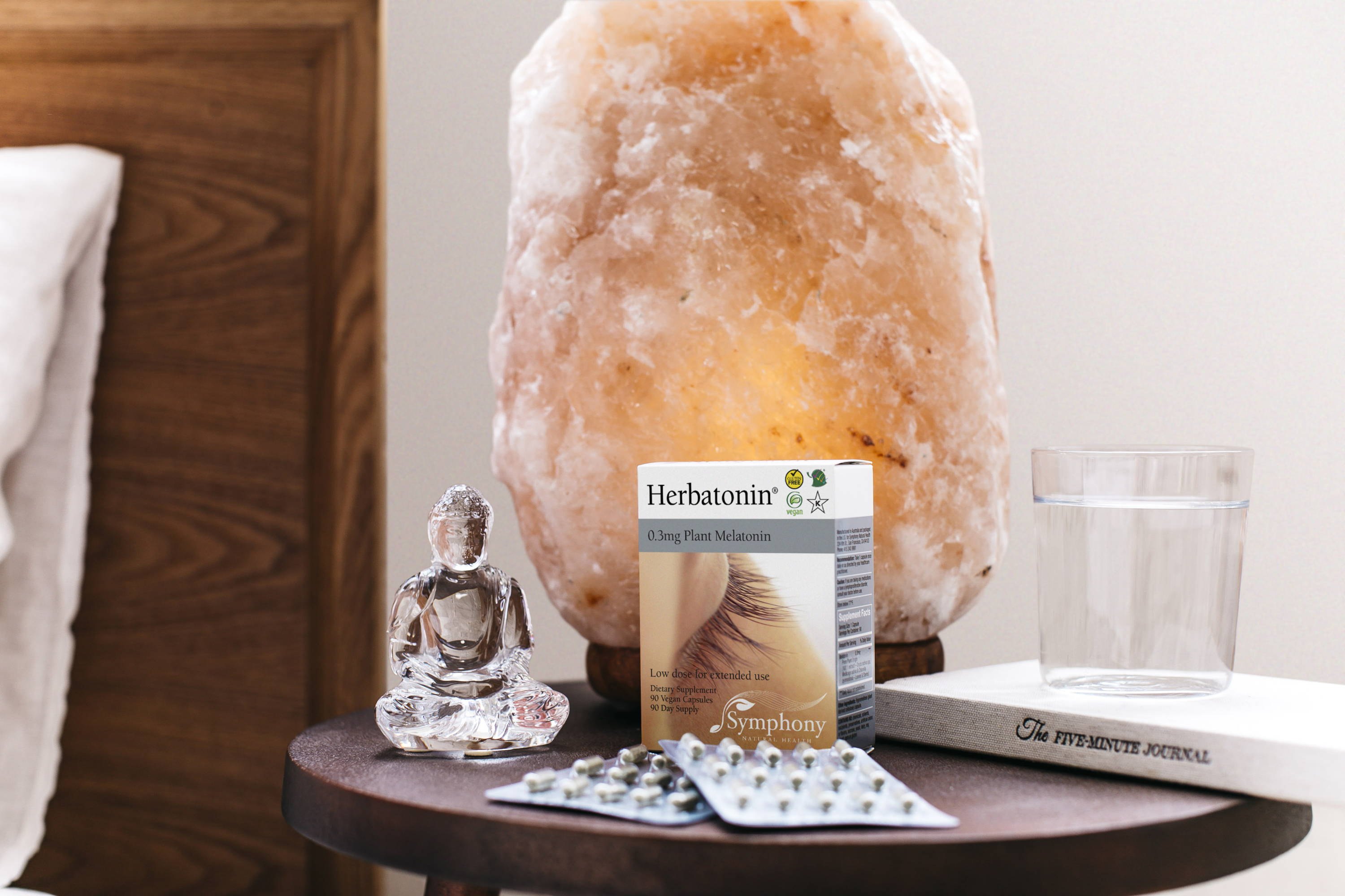 White bedding and brown wooden headboard next to brown wooden table with Herbatonin box and two blister packs, crystal figurine, himalayan crystal salt lamp, glass of water resting on book titled The Five-Minute Journal 