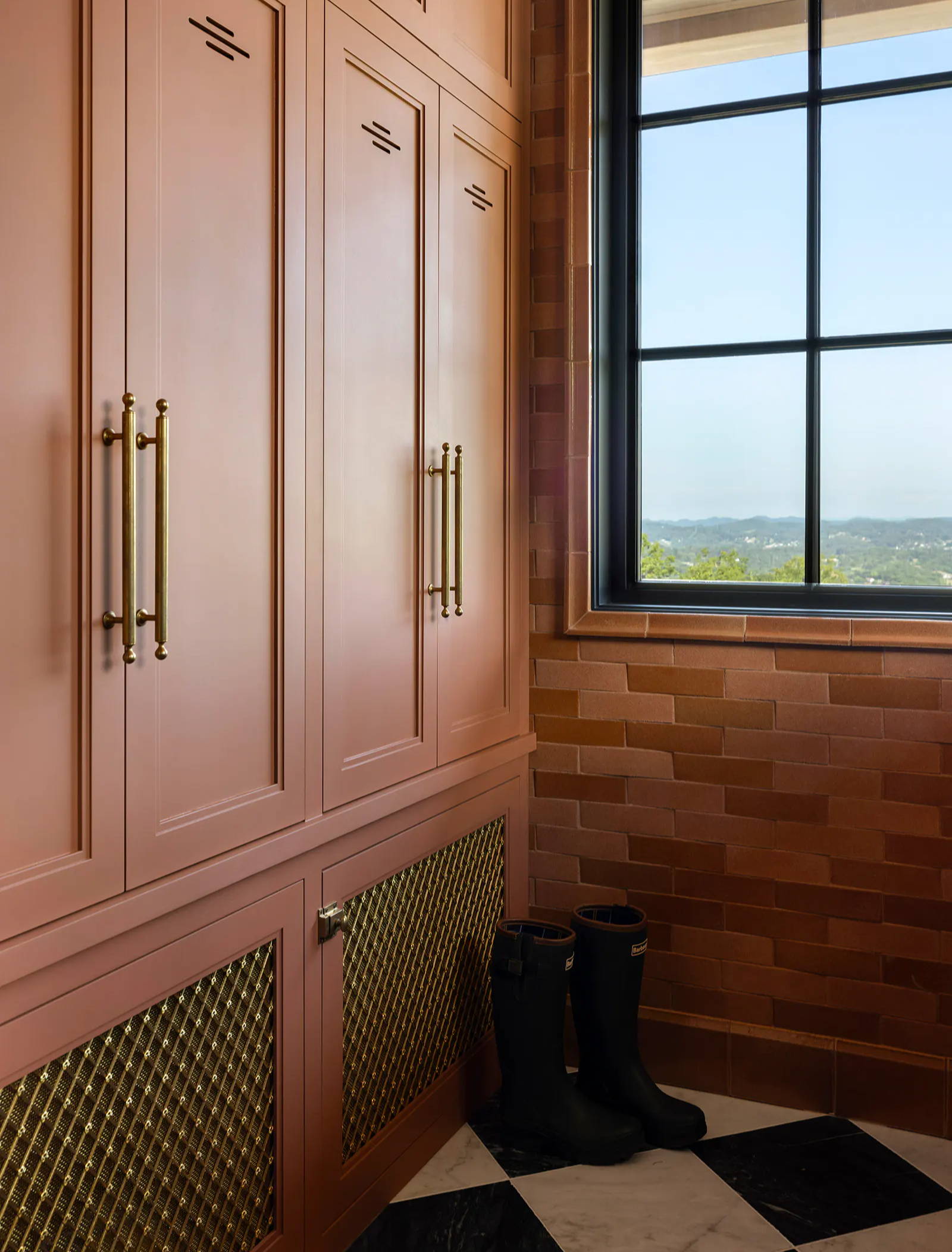 Colourful mudroom design by Well By Design with salmon pink cabinetry, brass hardware, chequered tiled flooring and exposed brick