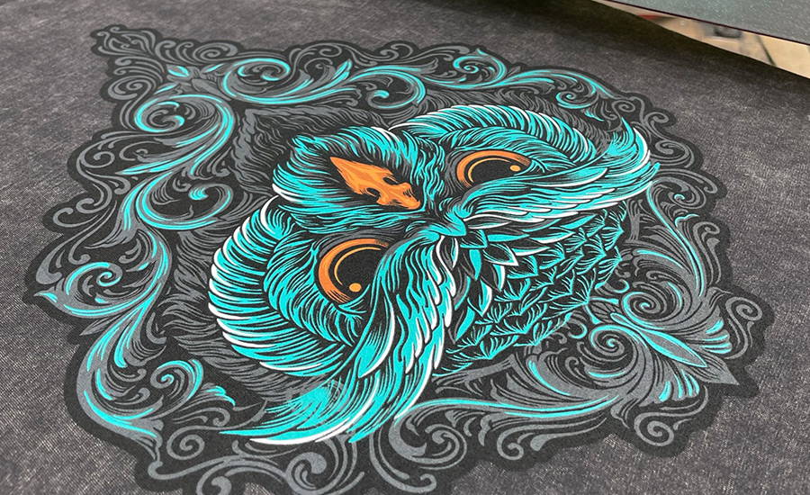A spot colour screen print design of an owls head inside a decorative filigree diamond frame on a distressed grey shirt. The owls feathers are predominantly aqua marine with white highlights, grey shadows, and black detailing. The beak and eyes have been printed in two bright oranges to give a popping contrast effect.