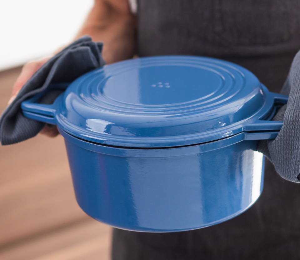 A chef carries a Blue Misen Dutch Oven covered with a Grill Lid easily, holding its wide handles with blue kitchen towels.