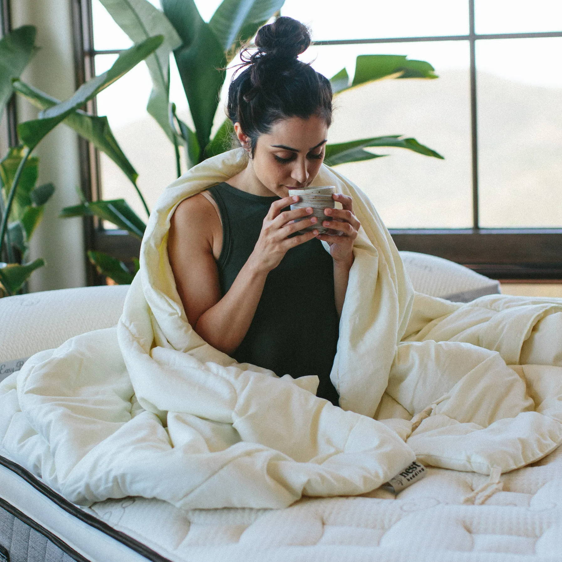 a woman sitting in bed sipping tea while wrapped in the nest bedding washable wool comforter