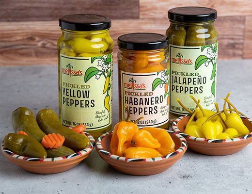 Jars of pickled peppers with bowls of assorted peppers on a table.