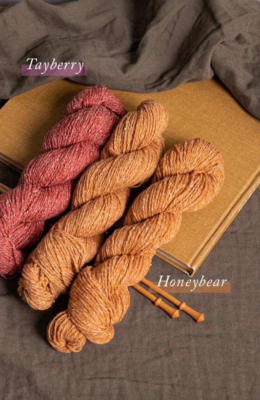 Three skeins of Dapple, two in color Honeybear and one in color Tayberry lie in a row on top of a linen covered book and a pair of knitting needles.