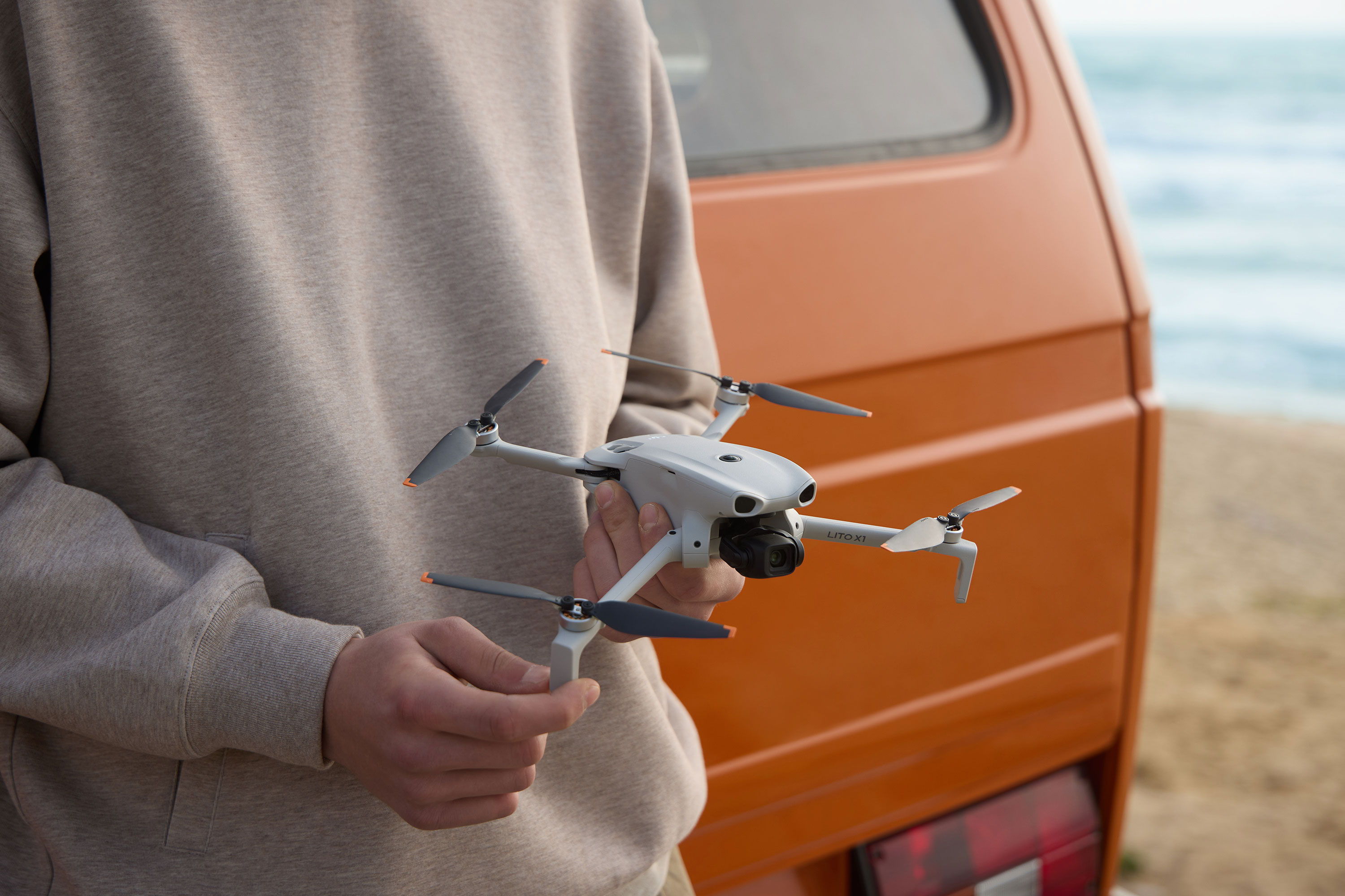 Person holding a DJI Lito X1 drone near an orange van by the beach.