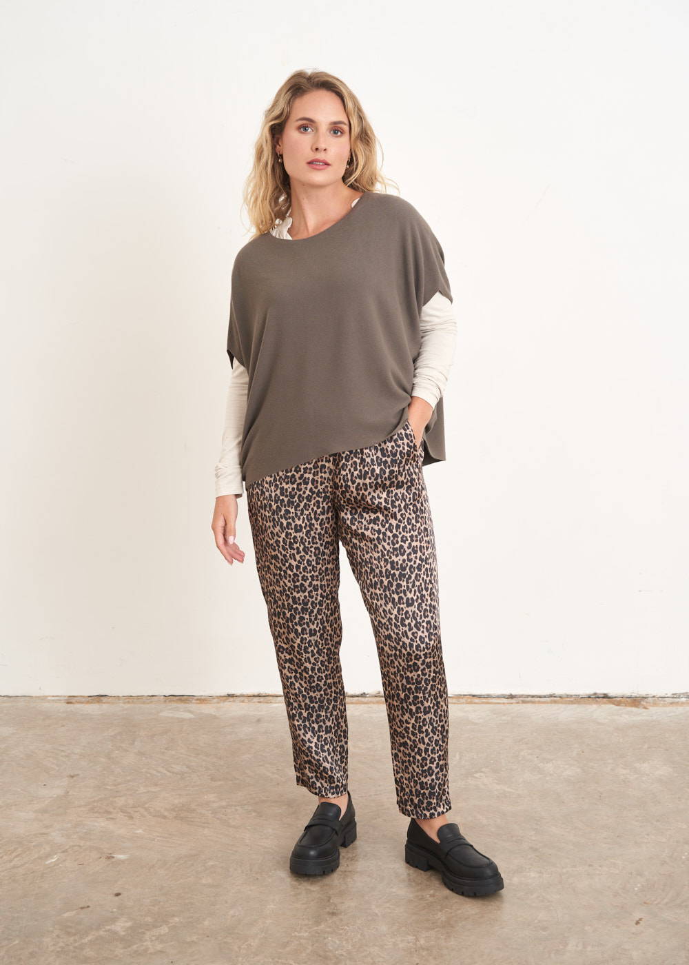 A model wearing a pair of golden leopard print trousers with a dark stone sleeveless blouse over a white top and with black loafers