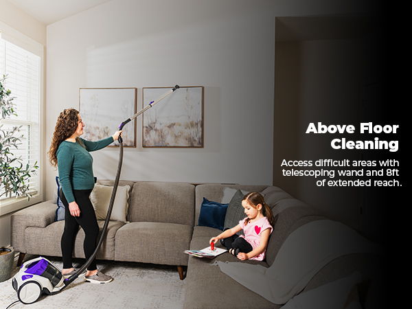 Woman cleaning above a couch with a vacuum; child playing nearby.