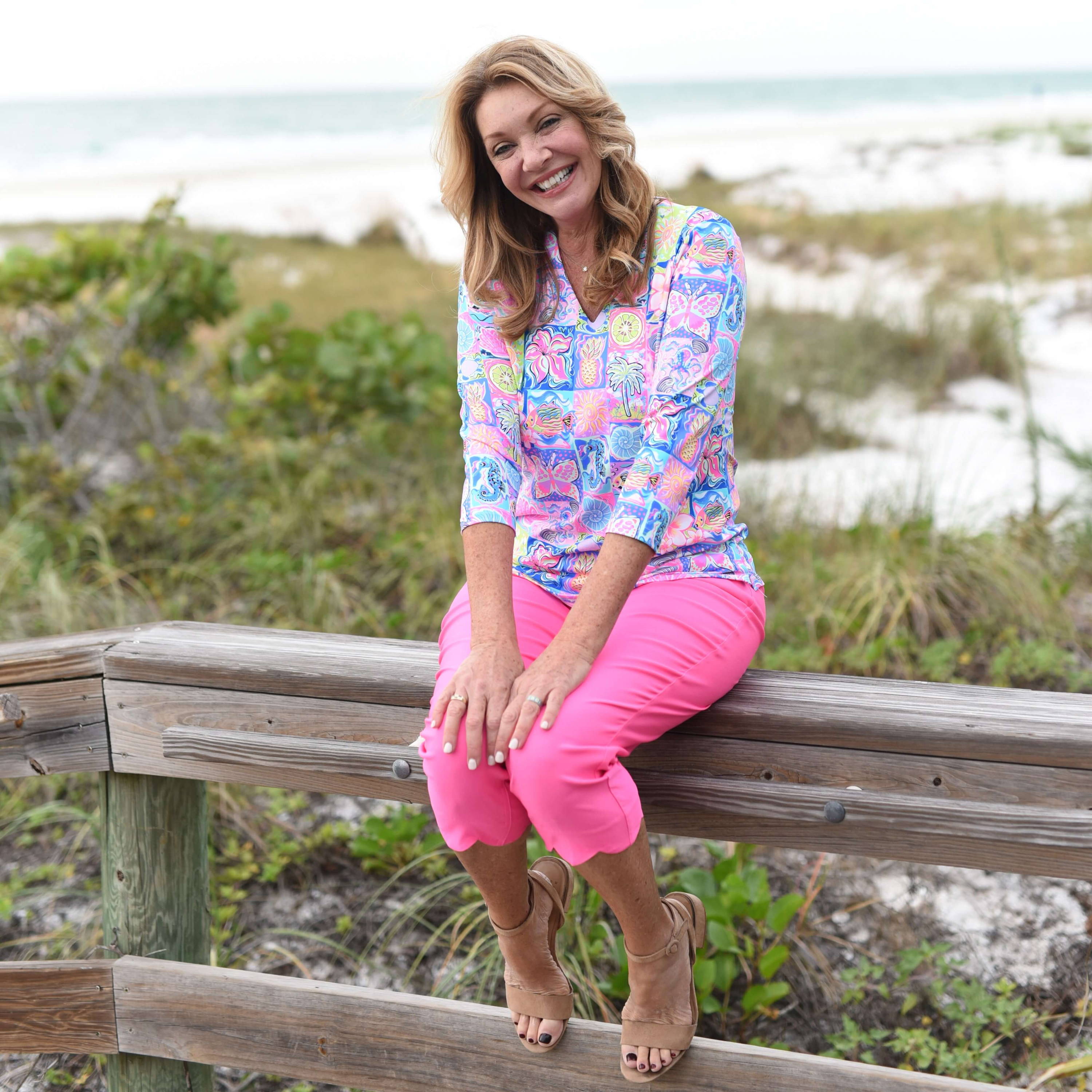 A smiling blonde woman wearing a printed top and pink capri pants sits on the ledge of a wooden boardwalk at the beach
