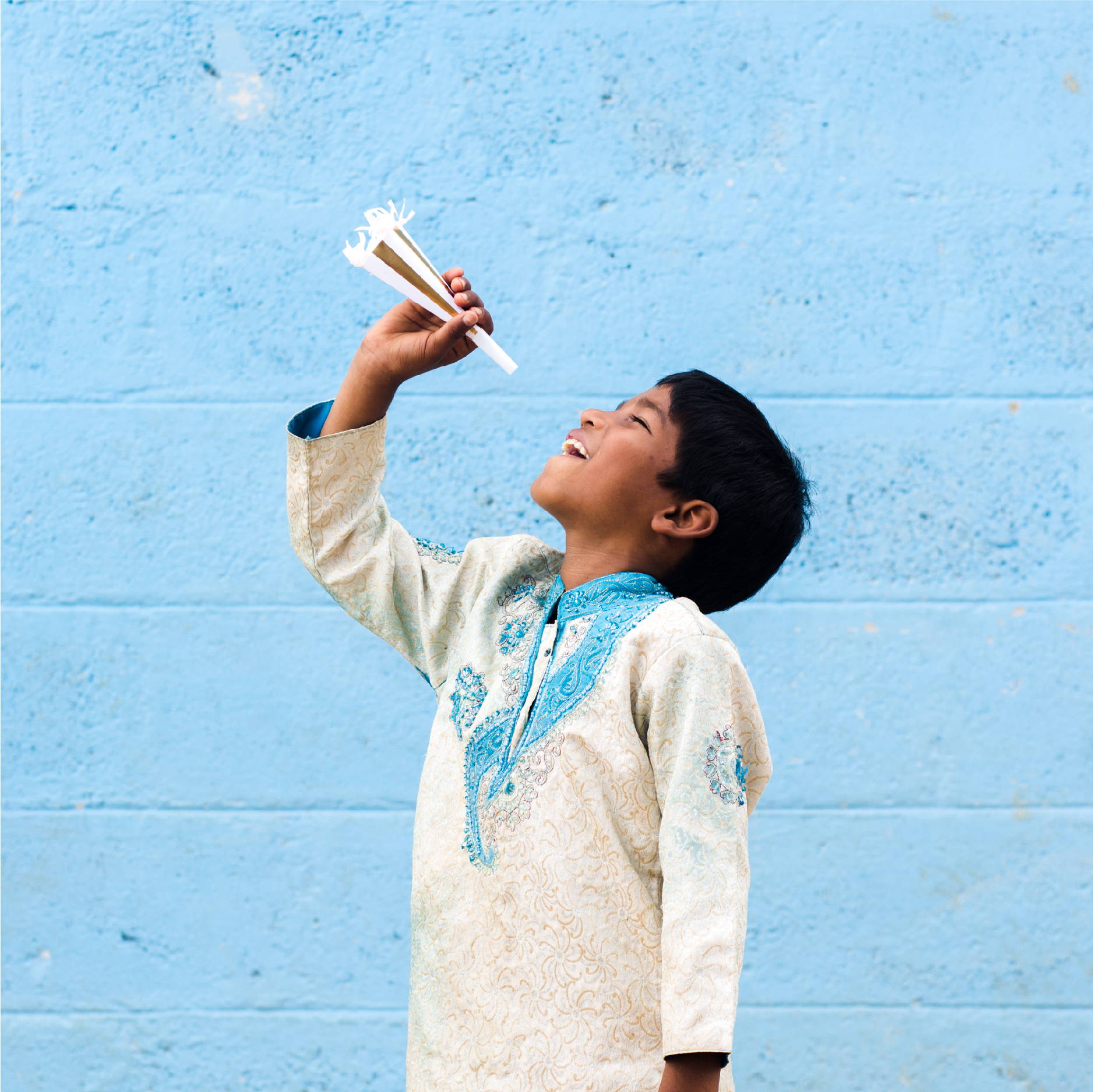 An Indian boy wearing traditional Indian clothing poses with a party horn in front of a blue wall with the look of pure joy 