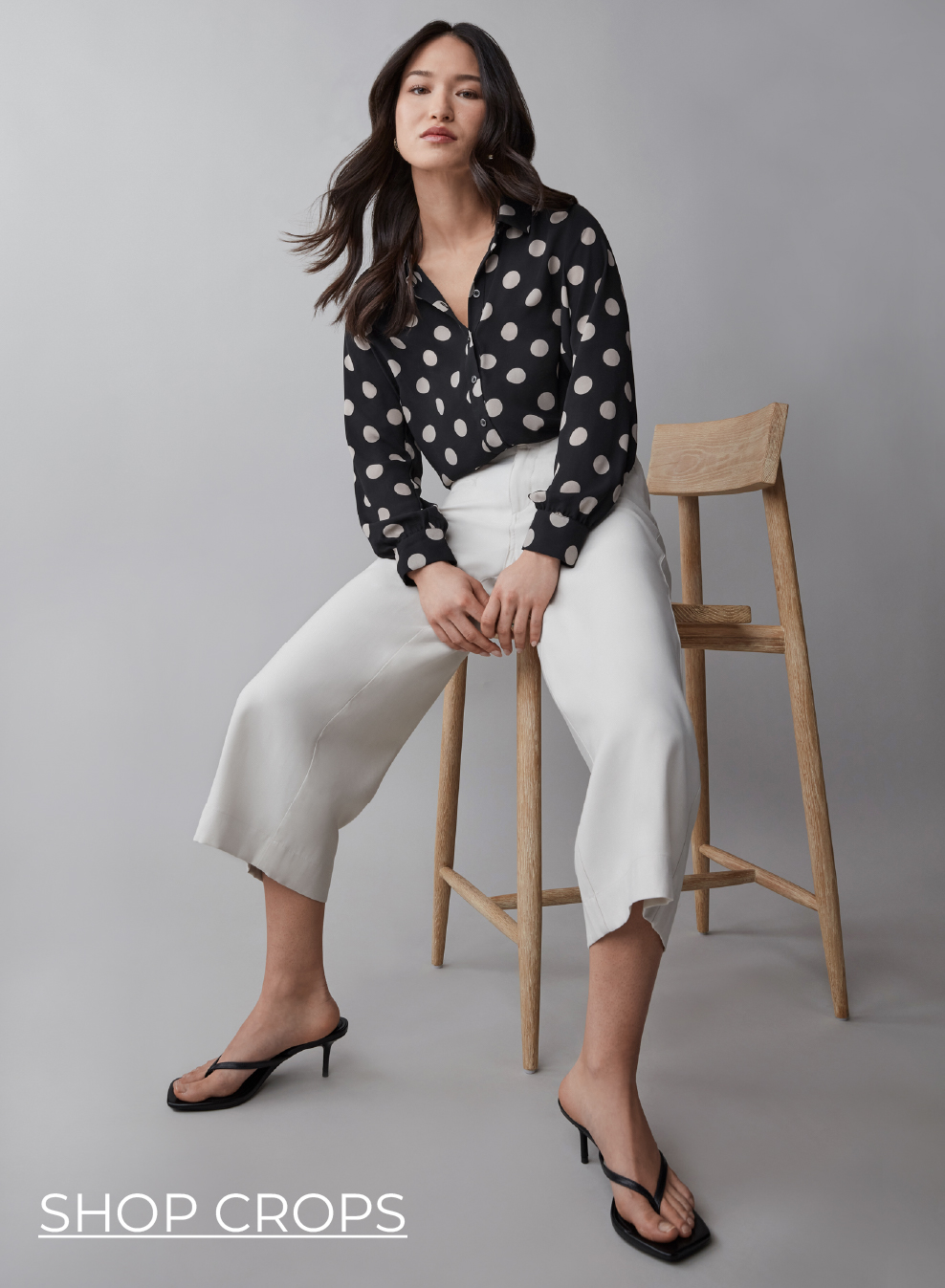 Model sitting on a stool, wearing a polka dot shirt and white pants.