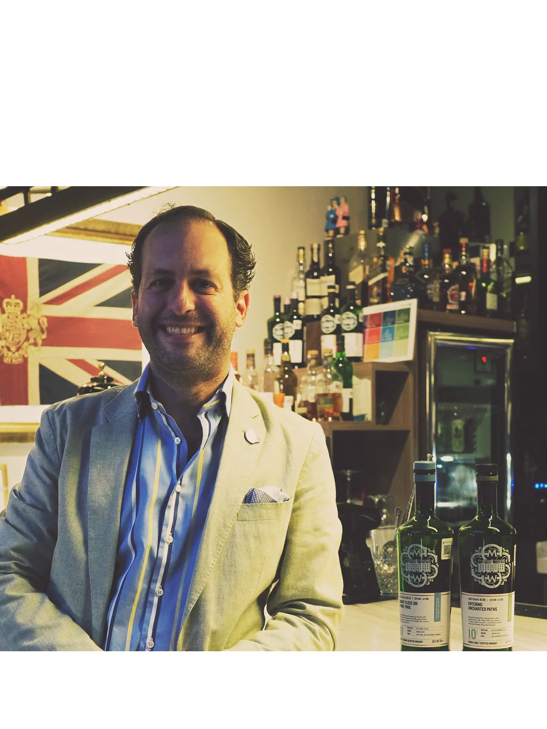Smiling man in a blazer stands beside bottles in a bar.