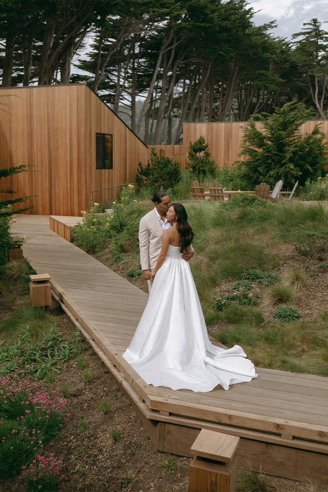 Couple embracing on a wooden path, surrounded by greenery and modern architecture.