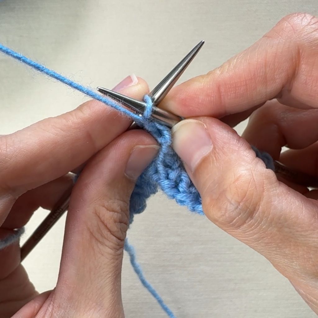 Detailfoto, wie eine linke Masche gestrickt wird. Die rechte Stricknadel sticht in die linke Masche von oben ein.