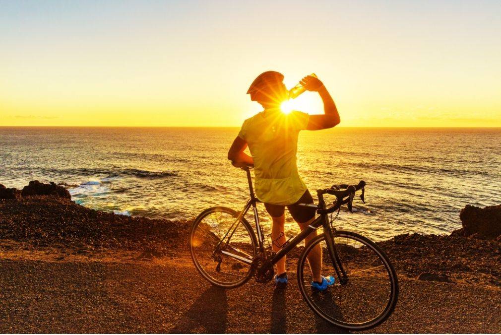 Männlicher Biker trinkt aus einer Wasserflasche, während die Sonne über dem Wasser untergeht