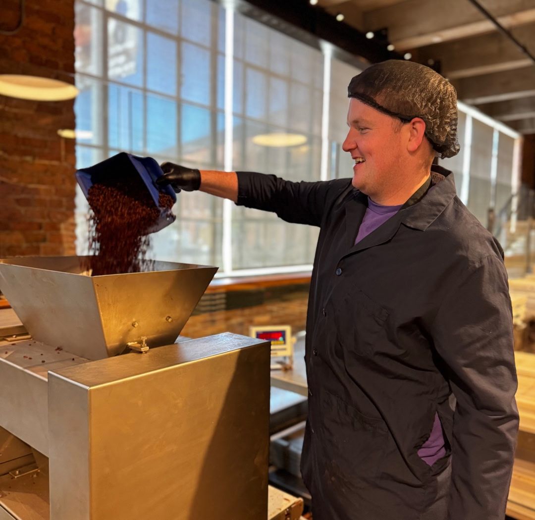 Person pouring coffee beans into a grinder in a bright, industrial setting.