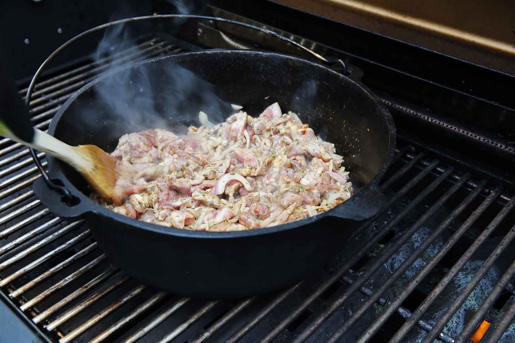 Cooking meat in a cast iron pot on a grill with steam rising.