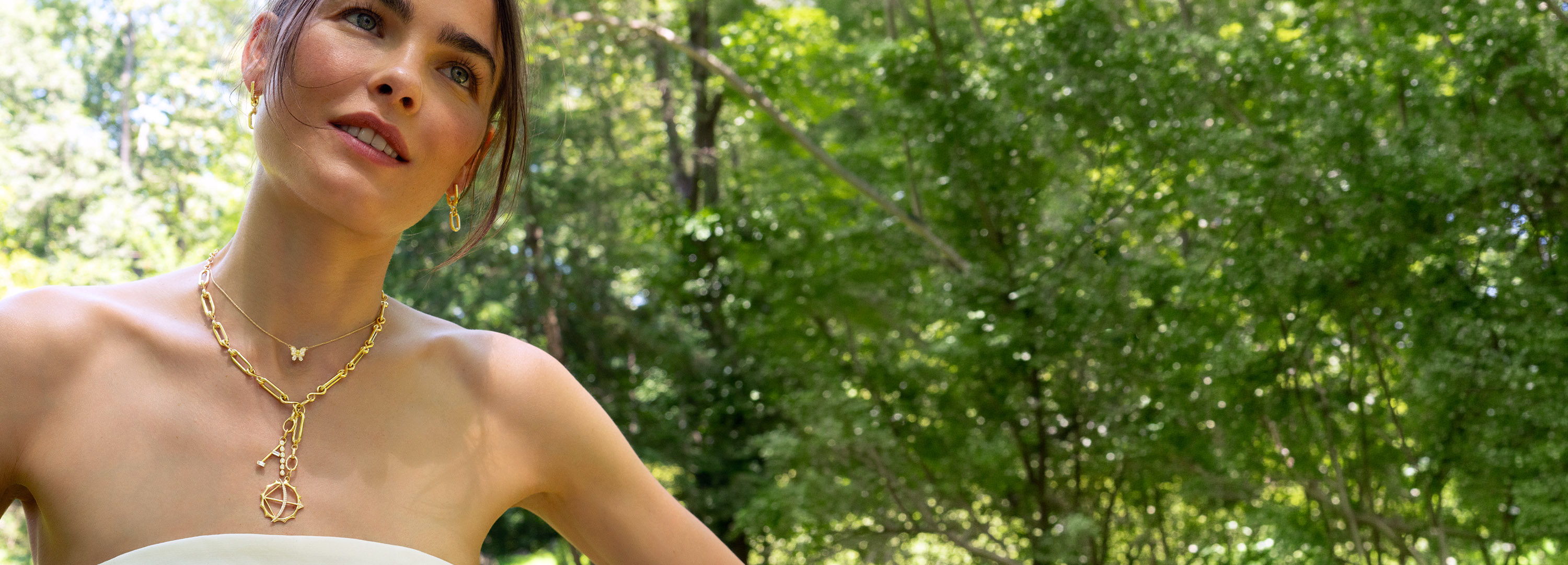 A woman in a white dress stands outdoors among green trees.