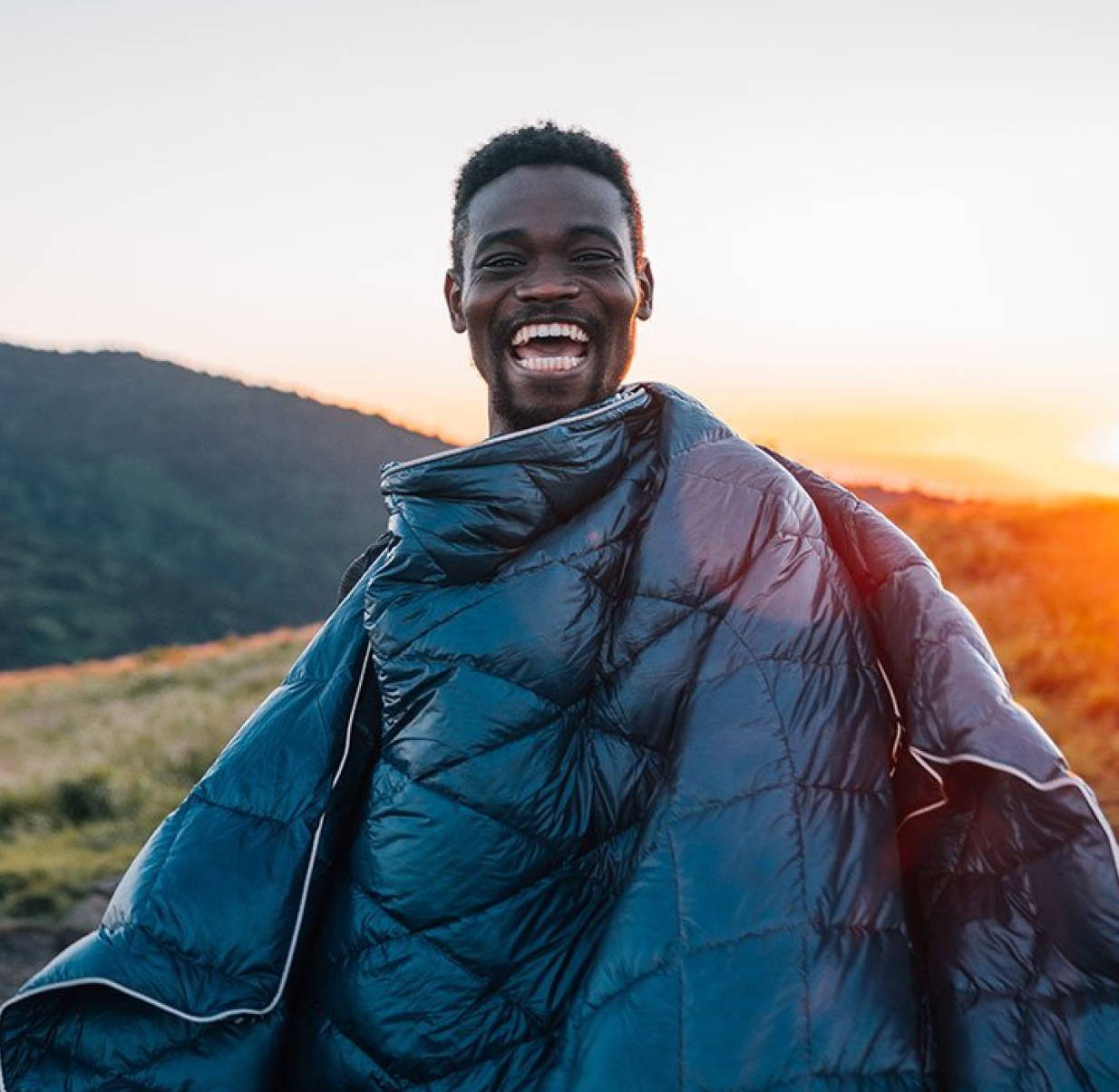 Man wearing a Rumpl Blanket as a cape and smiling at sunset
