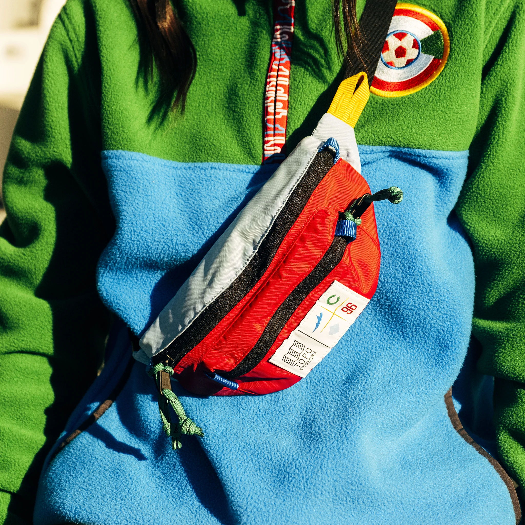 Person wearing a green and blue fleece with a red and gray pouch bag.