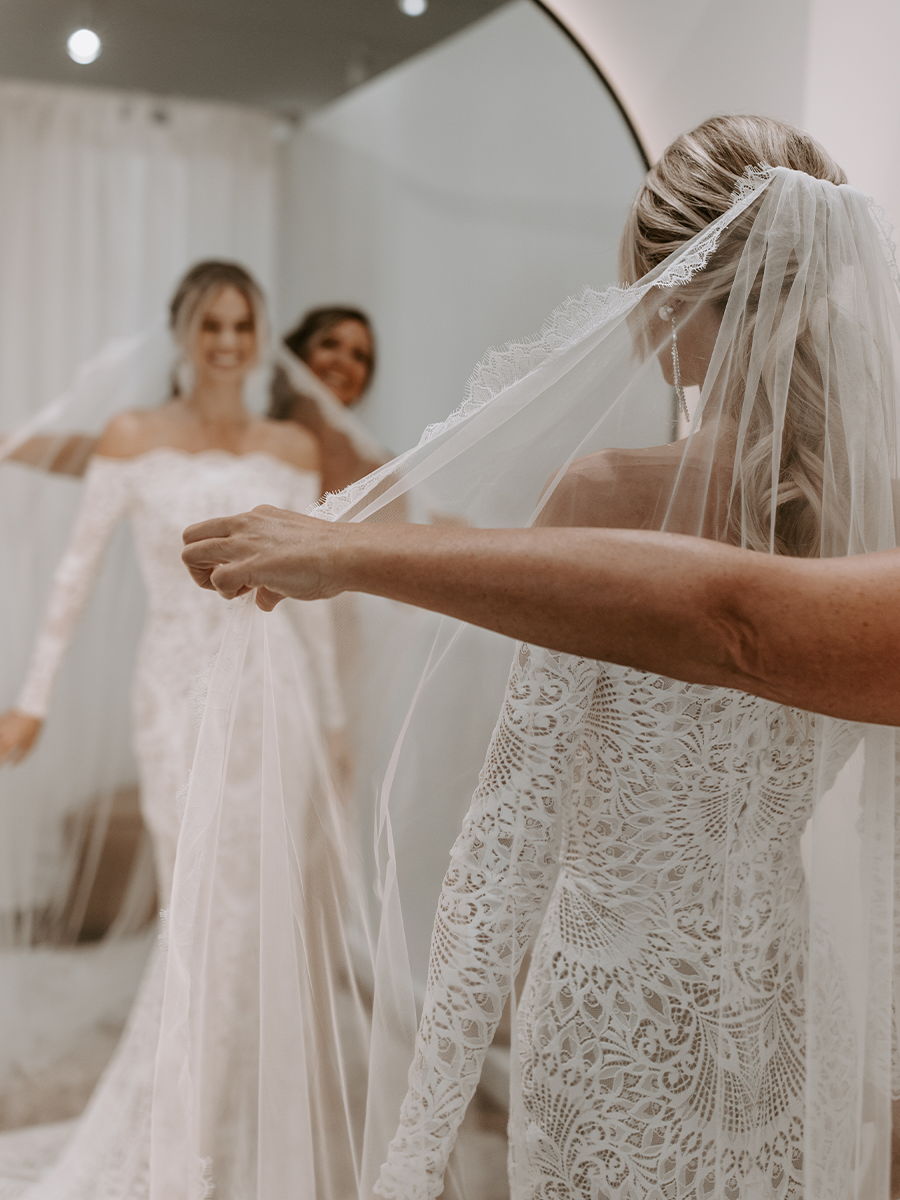 Bride trying on wedding dress with friends, adjusting veil in mirror reflection.