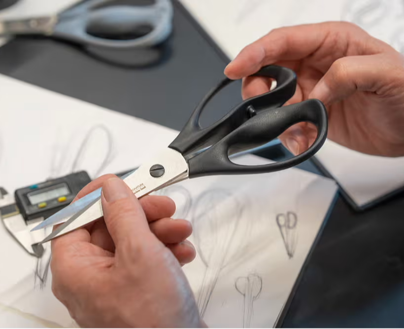 Person holding Signature Scissors over sketch papers on a table.