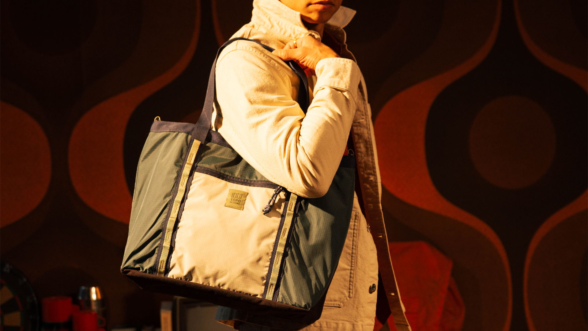 Person wearing a light jacket, holding a tote bag against a patterned background.